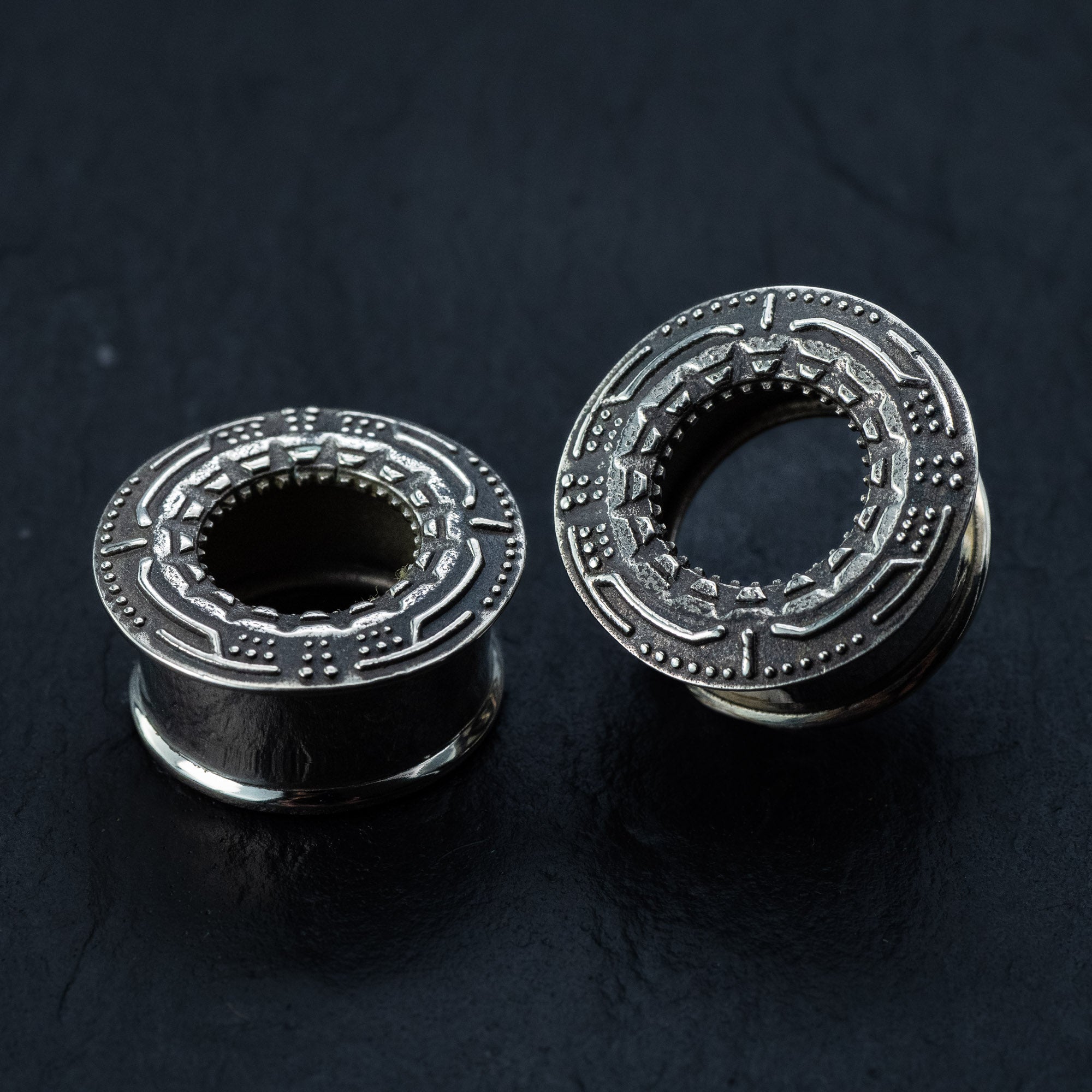 Two TERA Futuristic Sci Fi Flesh Tunnels from Kaya Movement, crafted in silver brass and featuring intricate geometric designs etched on their outer surfaces, rest on a dark, textured background. One tunnel stands upright, showcasing the detailed pattern around its hollow center, while the other lies on its side, highlighting its cylindrical shape—ideal for stretched ears.