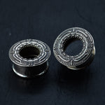Two TERA Futuristic Sci Fi Flesh Tunnels from Kaya Movement, crafted in silver brass and featuring intricate geometric designs etched on their outer surfaces, rest on a dark, textured background. One tunnel stands upright, showcasing the detailed pattern around its hollow center, while the other lies on its side, highlighting its cylindrical shape—ideal for stretched ears.