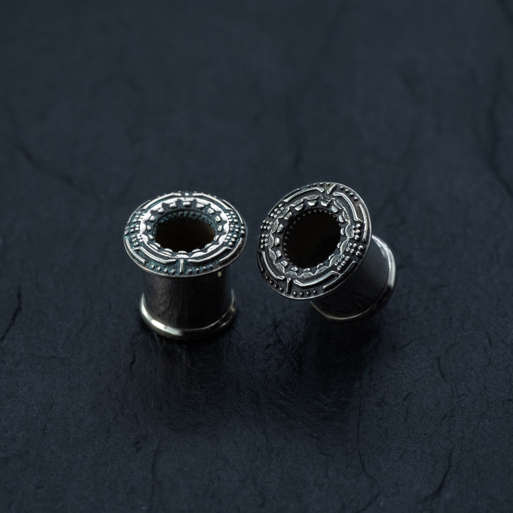 Two intricately designed, unisex TERA Futuristic Sci Fi Flesh Tunnels in Silver Brass by Kaya Movement are positioned on a dark, textured surface. The gauges range from 8mm to 20mm and have hollow centers with decorative rims featuring detailed patterns, presenting a polished, shiny appearance—perfect for those with stretched ears seeking futuristic body jewelry.