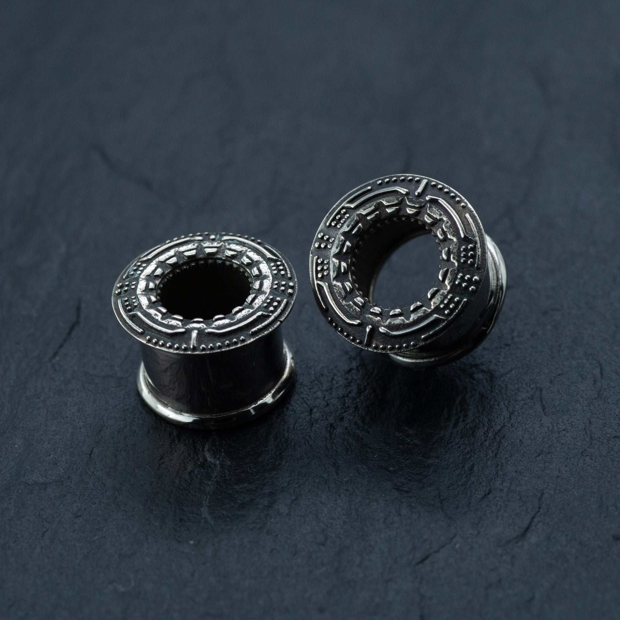 A pair of TERA Futuristic Sci Fi Flesh Tunnels by Kaya Movement, crafted from silver brass and available in sizes ranging from 8mm to 20mm, lie on a dark, textured surface. The unisex ear tunnels feature intricate circular geometric engravings and hollow centers. Positioned at different angles, they showcase their detailed craftsmanship—ideal for those with stretched ears seeking a touch of futuristic body jewelry.