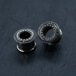 A pair of TERA Futuristic Sci Fi Flesh Tunnels by Kaya Movement, crafted from silver brass and available in sizes ranging from 8mm to 20mm, lie on a dark, textured surface. The unisex ear tunnels feature intricate circular geometric engravings and hollow centers. Positioned at different angles, they showcase their detailed craftsmanship—ideal for those with stretched ears seeking a touch of futuristic body jewelry.