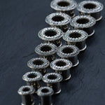 A set of twenty silver brass TERA Futuristic Sci Fi Flesh Tunnels, crafted by Kaya Movement, is arranged on a dark surface. The tunnels, ranging from 8mm to 20mm in size, are lined up diagonally to create a pattern reminiscent of futuristic body jewelry. The lighting highlights their metallic texture and detailed craftsmanship.