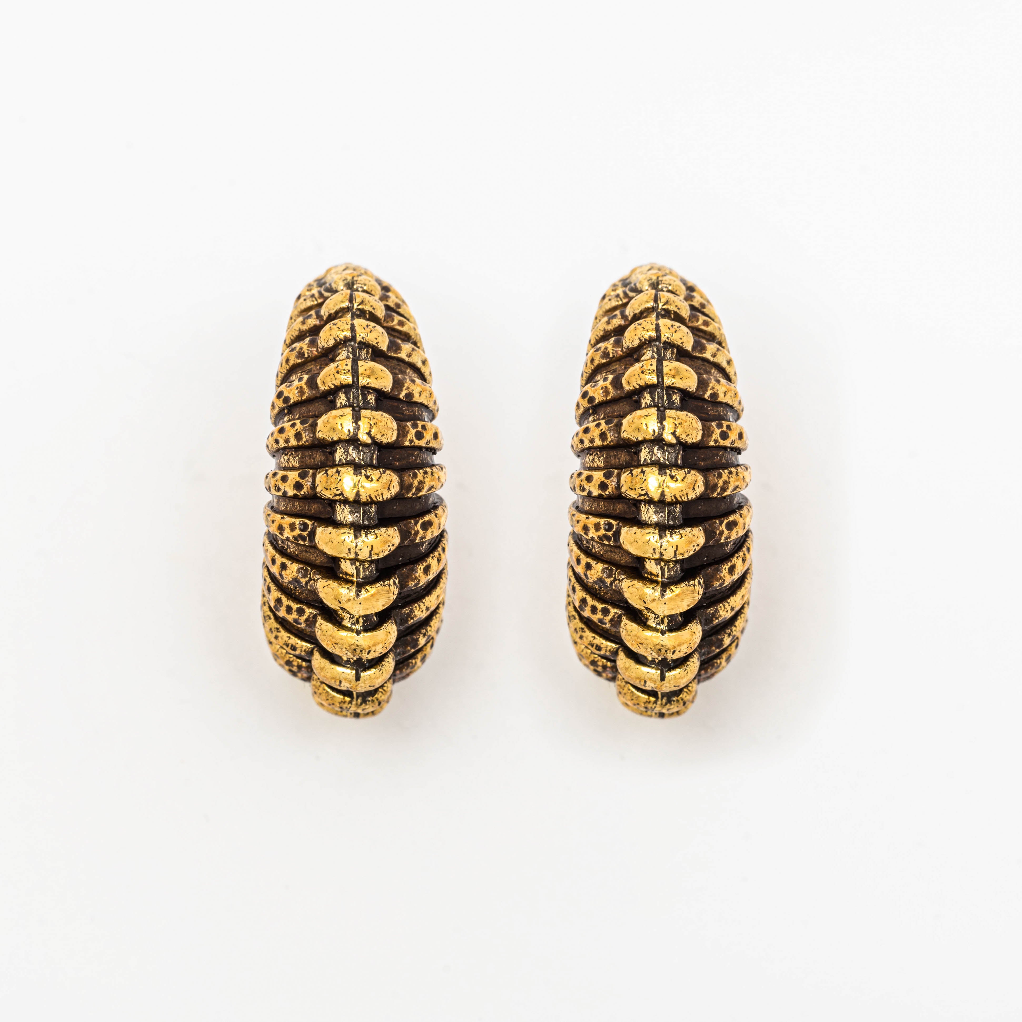 A close-up image featuring two decorative TRILOBITE Large Biomechanical Clicker Ear Weights in Gold Brass by Kaya Movement. The ear weights display a cylindrical, segmented design with a ridged black and gold pattern, evoking the aesthetic of ancient tribal gauge earrings. They are arranged vertically side by side on a white background.