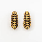 A close-up image featuring two decorative TRILOBITE Large Biomechanical Clicker Ear Weights in Gold Brass by Kaya Movement. The ear weights display a cylindrical, segmented design with a ridged black and gold pattern, evoking the aesthetic of ancient tribal gauge earrings. They are arranged vertically side by side on a white background.