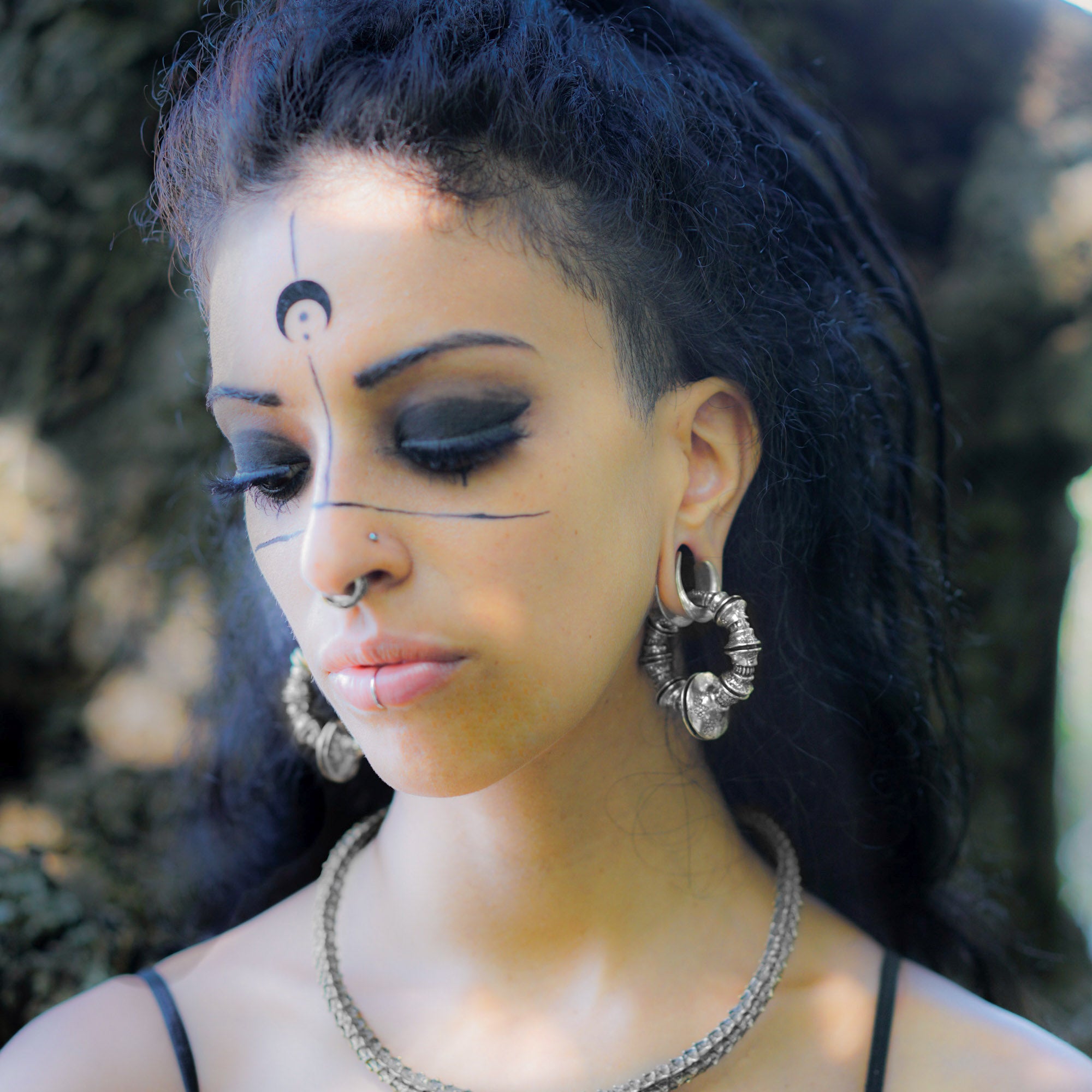 A person with dark hair and elaborate, silver jewelry poses outdoors. They have bold black eye makeup, intricate face paint with lines and a crescent moon, complemented by the Kaya Movement's TRAKARA Heavy Tribal Cyborg Clicker Ear Weights in Silver (2 gauge). The background suggests a natural, wooded environment.