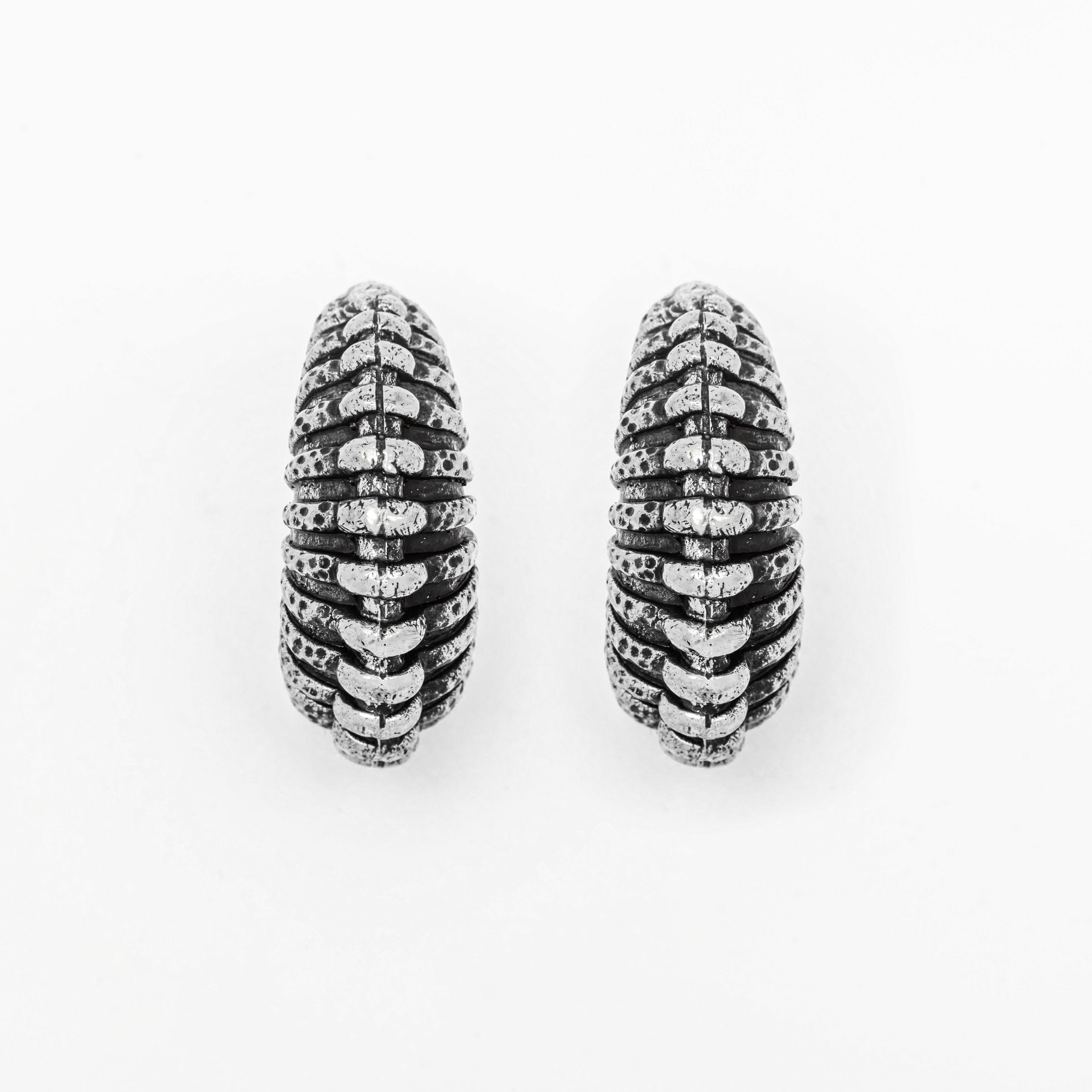 A pair of oxidized TRILOBITE Large Biomechanical Clicker Ear Weights in Silver Brass from Kaya Movement. Resembling post-apocalyptic body jewelry, the 0 gauge ear weights feature a unique pattern of horizontal ridges, creating a ribbed appearance. The edges are slightly darker, accentuating the intricate detailing against a plain white background.