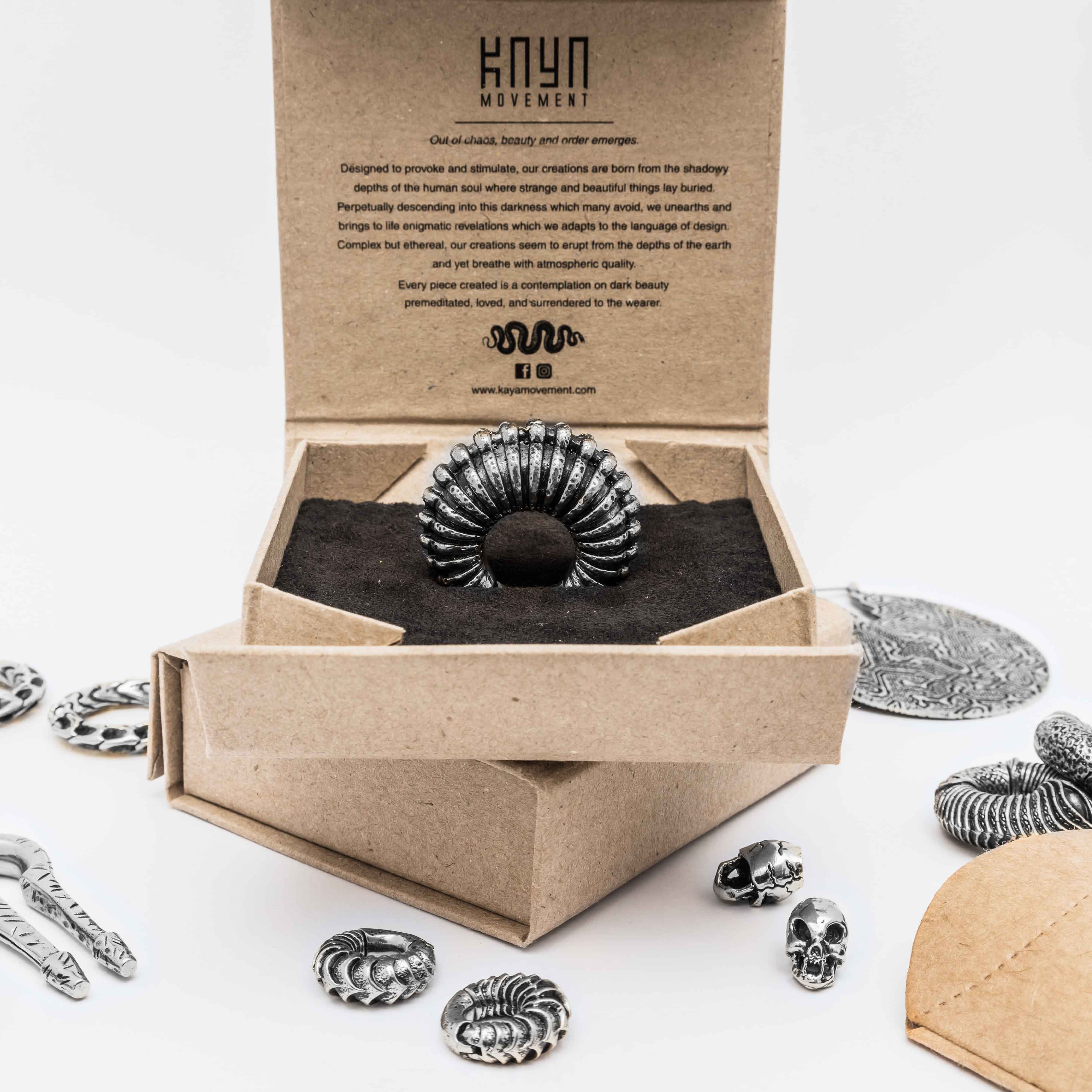 A decorative ring featuring a feather-like design sits on display in an open cardboard box with the "Kaya Movement" logo. The background showcases additional metallic jewelry, including TRAKARA Heavy Tribal Cyborg Clicker Ear Weights in Silver (2 gauge), a circular medallion, skull charms, and various ornate designs scattered around.