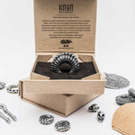 A decorative ring featuring a feather-like design sits on display in an open cardboard box with the "Kaya Movement" logo. The background showcases additional metallic jewelry, including TRAKARA Heavy Tribal Cyborg Clicker Ear Weights in Silver (2 gauge), a circular medallion, skull charms, and various ornate designs scattered around.