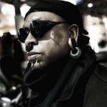 A person with dark skin wearing futuristic sunglasses, a black headscarf, and a dark jacket can be seen. They have various piercings, including a septum ring, lip piercings, and the TRILOBITE Large Biomechanical Clicker Ear Weights in Silver Brass by Kaya Movement in their ears. The background is blurred, providing a stark contrast to their detailed appearance.
