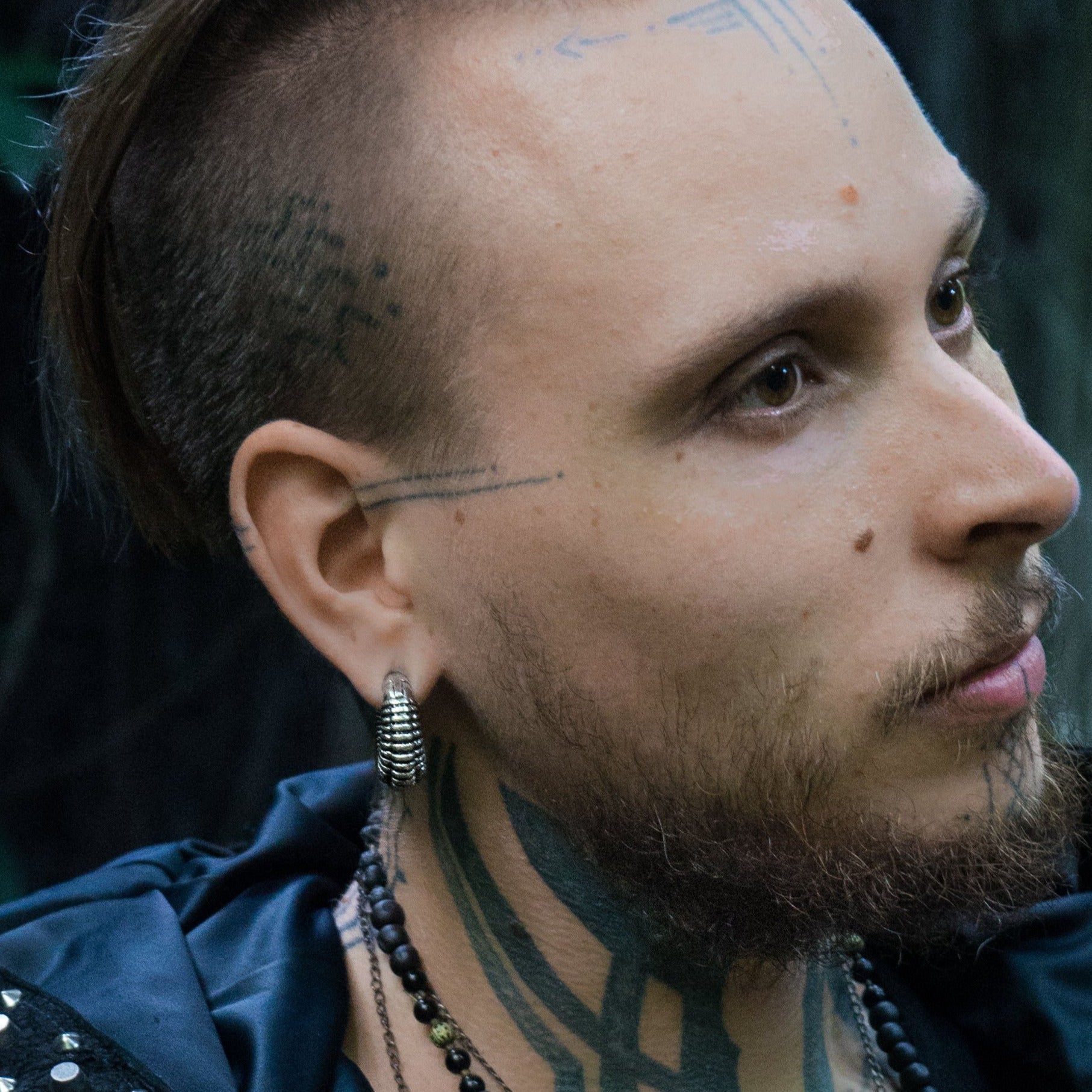 A person with a side-shaved hairstyle, intricate tattoos on their face and neck, and wearing TRILOBITE Small Biomechanical Clicker Ear Weights in Silver Brass (2 gauge) by Kaya Movement and beaded necklaces looks thoughtfully into the distance. The background appears to be an outdoor setting with blurred, dark foliage.