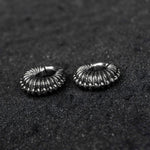 Close-up image of two TRILOBITE Large Biomechanical Clicker Ear Weights in Silver Brass from Kaya Movement, resting on a dark, coarse surface. The ribbed designs give these 0 gauge ear weights a distinctive, intricate appearance reminiscent of post-apocalyptic body jewelry.