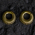 A pair of TRILOBITE Small Biomechanical Clicker Ear Weights in Gold Brass by Kaya Movement rests on a textured black surface. The ear weights, designed in a 2 gauge size, feature a vintage appearance with their distinct ridged design and circular shape that evokes tribal gauge earrings. The black background contrasts with the shiny gold brass, emphasizing the intricate details.