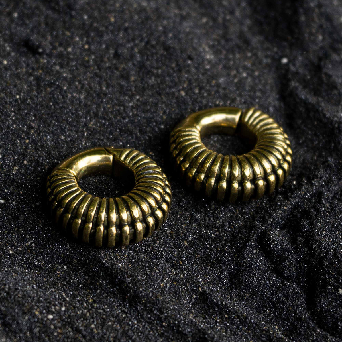 A pair of TRILOBITE Small Biomechanical Clicker Ear Weights in Gold Brass by Kaya Movement lie on a textured dark surface, possibly sand or coarse fabric. The surface's rough texture contrasts with the smooth, shiny finish of the bold, intricately designed 2 gauge tribal earrings.