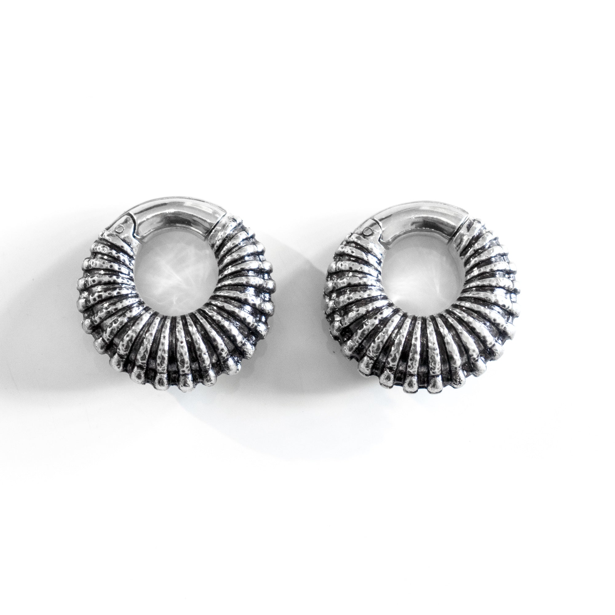 Introducing the TRILOBITE Large Biomechanical Clicker Ear Weights in Silver Brass by Kaya Movement. These 0 gauge earrings feature a round, ribbed, ridged texture with a polished metallic finish. Resembling post-apocalyptic body jewelry, they are elegantly displayed on a white background.

