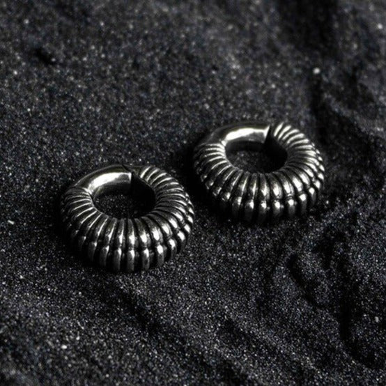 A pair of TRILOBITE Small Biomechanical Clicker Ear Weights in Silver Brass, 2 gauge by Kaya Movement, are placed on a textured, dark surface resembling granular sand or gravel. The earrings have a shiny finish, reflecting light subtly.