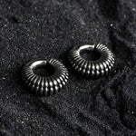 A pair of TRILOBITE Small Biomechanical Clicker Ear Weights in Silver Brass, 2 gauge by Kaya Movement, are placed on a textured, dark surface resembling granular sand or gravel. The earrings have a shiny finish, reflecting light subtly.