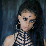 A person with long, curly black hair is wearing Quetzalcoatl Oversized Snake Spine Hoop Earrings in Silver White Brass by Kaya Movement and a black harness outfit. They have face markings, including lines and a crescent moon symbol on their forehead. The background is blurry, suggesting an indoor setting.