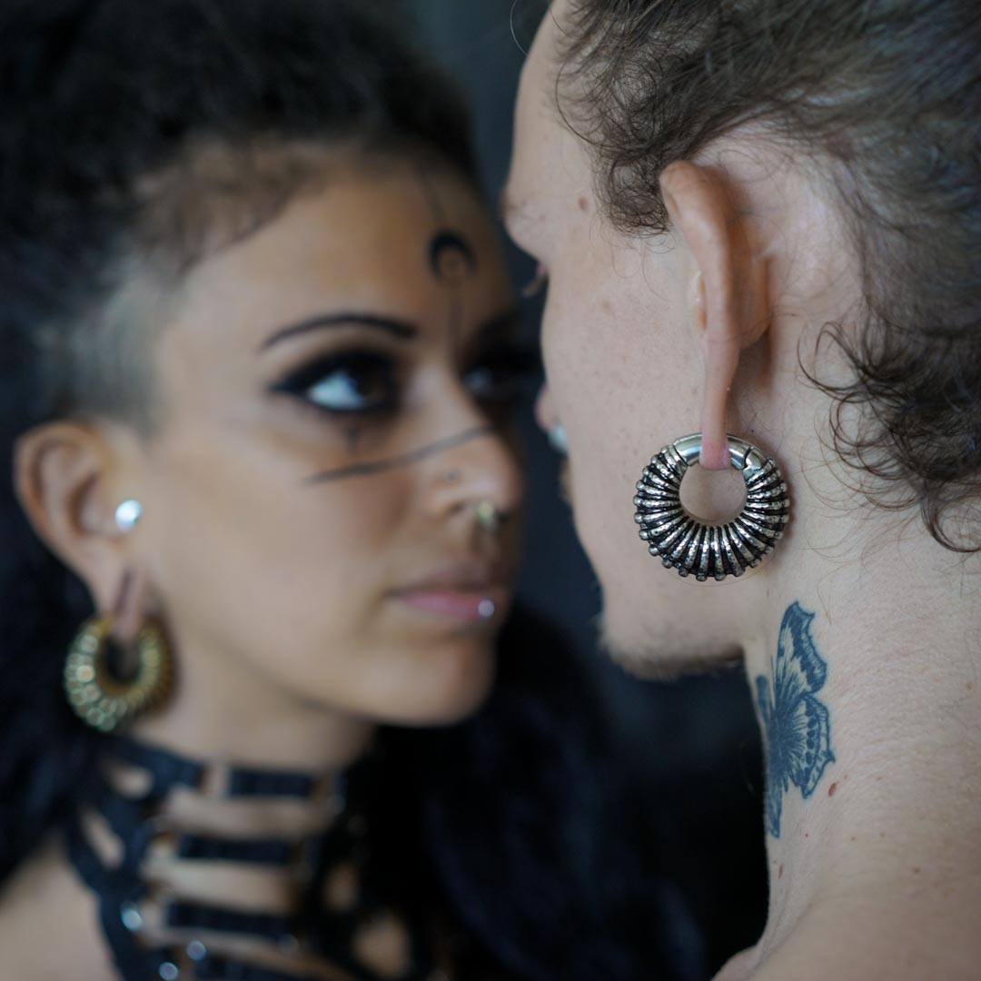 Two individuals with striking facial and ear piercings are facing each other. One, a woman with intricate facial tattoos, black eye makeup, and large post-apocalyptic body jewelry hoop earrings, gazes at the other individual, who has a neck tattoo and wears Kaya Movement’s TRILOBITE Large Biomechanical Clicker Ear Weights in Silver Brass | 0 gauge.