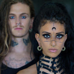 A close-up of two individuals with alternative styles. The person in the foreground has long black hair, facial piercings, face tattoos, and a black strappy outfit accented by TRILOBITE Small Biomechanical Clicker Ear Weights in Gold Brass from Kaya Movement. The person in the background has long hair, facial piercings, and tattoos on their neck and chest.