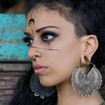 A person adorned with large circular earrings featuring an intricate design and the DARMA Biomechanical Clicker Hoop Septum Nose Ring in Gold by Kaya Movement poses thoughtfully. They sport elaborate black face paint with a crescent moon on their forehead and lines extending downward from their eyes, set against a blurred background.