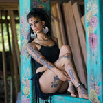 A woman with tattoos is sitting on a turquoise and floral-painted wooden column. She has long, curly hair and is wearing large, intricate CIRCUIT Oversized Visionary Disc Hoop Earrings in Gold by Kaya Movement, a black crop top, and a black skirt. Her expression is calm as she looks directly at the camera.