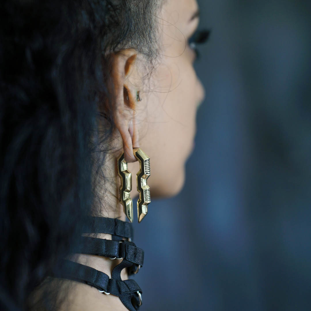 A person with long, dark hair shown from the side wears Kaya Movement's large, intricate TECHRE Futuristic Sci Fi Hexagon Keyhole Ear Weights in Gold, designed for a 0 gauge. The individual's neck is adorned with a black choker-style accessory. The background is blurred and dark, emphasizing the focus on the earrings and necklace.
