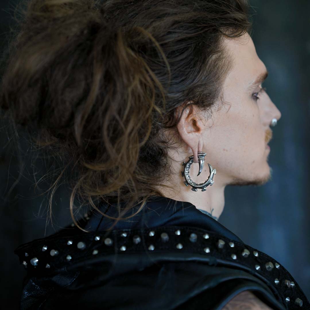 Profile of a person with long, messy hair tied back, wearing a studded black leather jacket and the GIGER Biomechanical Open Ended Hoop Ear Weights in Silver 925 from Kaya Movement. They have a nose piercing and their face is turned to the right, showing a calm expression against a dark background.
