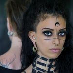 A woman with intricate facial markings and piercings stares intently forward. She has dark makeup and wears a black choker with metal rings. Another person, adorned with Kaya Movement's TRILOBITE Small Biomechanical Clicker Ear Weights in Gold Brass on their tattooed shoulder, stands in the background facing away. The scene is artistic and moody.