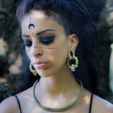 A woman with dark eye makeup and black facial markings, including a crescent moon on her forehead, stands outdoors. She has large TRAKARA Heavy Tribal Cyborg Clicker Ear Weights in Gold (2 gauge) by Kaya Movement and a nose ring, and wears a braided necklace. Her hair is partially shaved on one side, exuding a post-apocalyptic body jewelry vibe.
