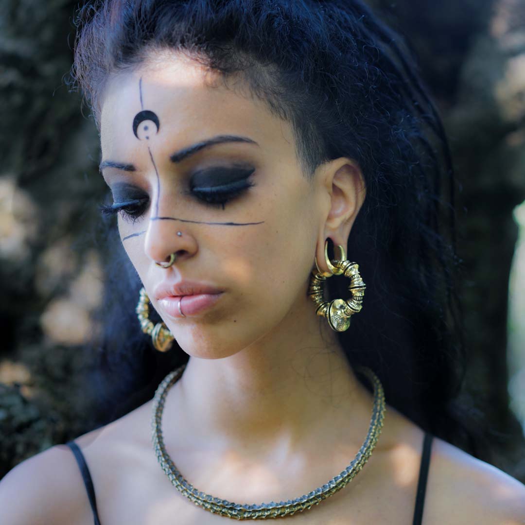 A woman with dark eye makeup and black facial markings, including a crescent moon on her forehead, stands outdoors. She has large TRAKARA Heavy Tribal Cyborg Clicker Ear Weights in Gold (2 gauge) by Kaya Movement and a nose ring, and wears a braided necklace. Her hair is partially shaved on one side, exuding a post-apocalyptic body jewelry vibe.