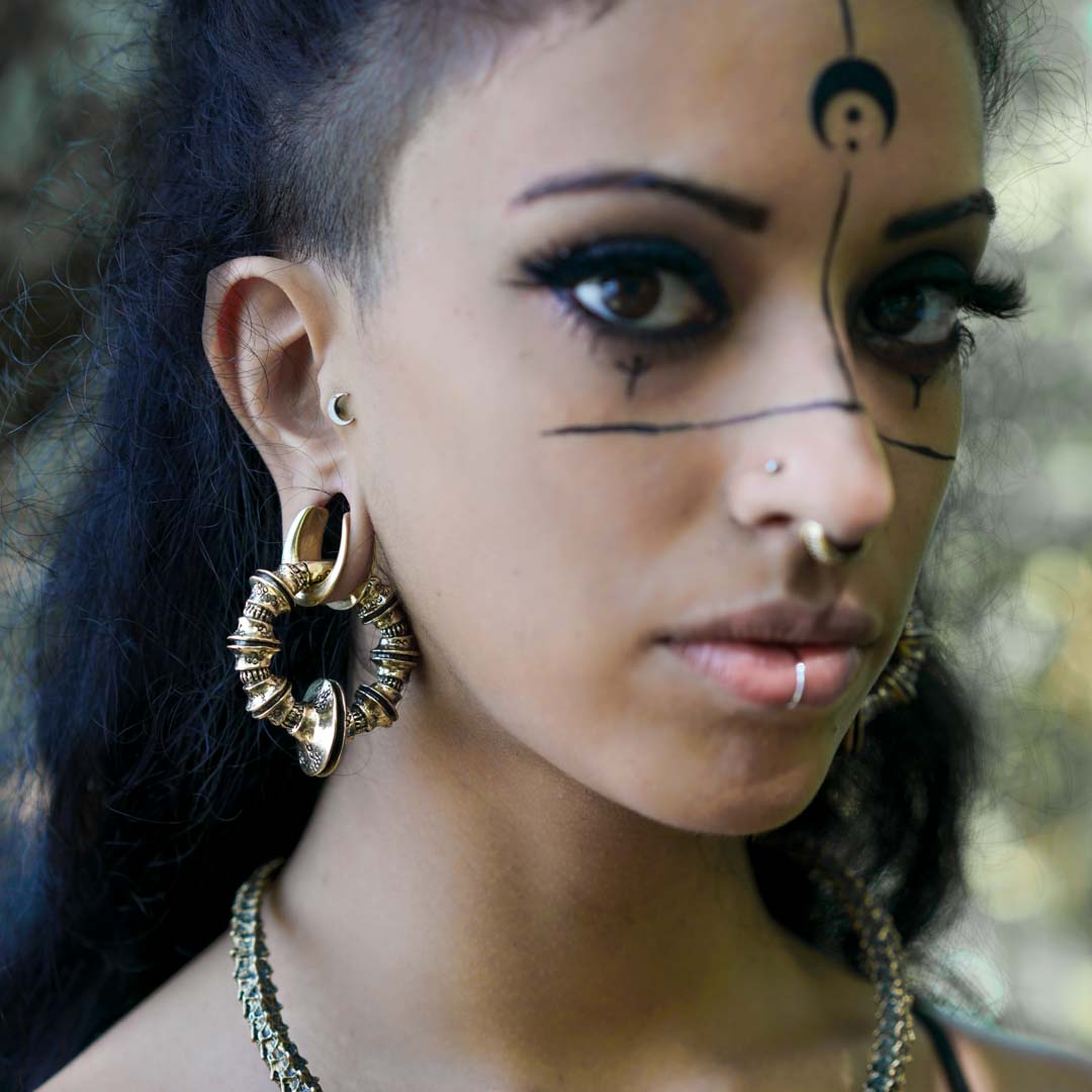 A person with intricate black face paint, including lines stretching from their eyes across their cheeks and a design on their forehead, is wearing large, ornate TRAKARA Heavy Tribal Cyborg Clicker Ear Weights in Gold (2 gauge) by Kaya Movement and post-apocalyptic body jewelry like a nose ring and a subtle lip ring. They have dark hair and are looking directly at the camera.