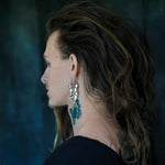 Side profile of a person with long, wavy hair wearing Kaya Movement's TECHRE Futuristic Hexagon Keyhole Ear Weights in Silver (0 gauge), reminiscent of post-apocalyptic body jewelry. The person showcases multiple tattoos, including one on the neck and another partially visible on the upper back. The background is dark and blurred.