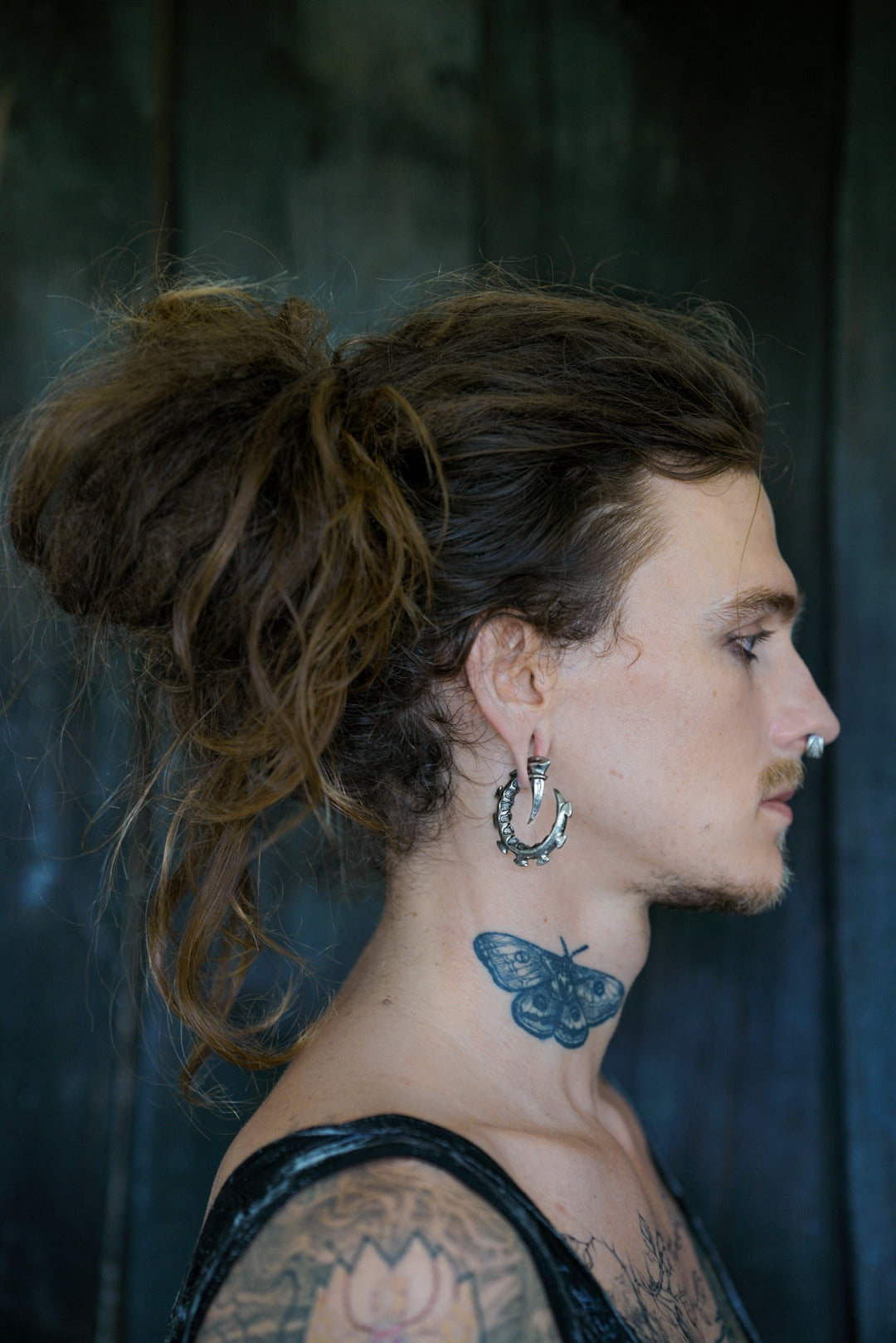 A person with long, curly brown hair in a messy bun, wearing Kaya Movement's GIGER Biomechanical Open Ended Hoop Ear Weights in Silver 925 (0 gauge) and post-apocalyptic body jewelry, has a side profile visible. They have a butterfly tattoo on their neck and a nose ring, displaying a patterned tattoo on their arm against a dark background.