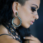 A close-up shot of a person with long dark hair and dramatic eye makeup, sporting a nose ring and two sci-fi statement earrings: the Quetzalcoatl Oversized Snake Spine Hoop Earrings by Kaya Movement in Silver White Brass. Wearing a harness-style accessory, they gently touch their hair, creating an edgy post-apocalyptic body jewelry vibe.