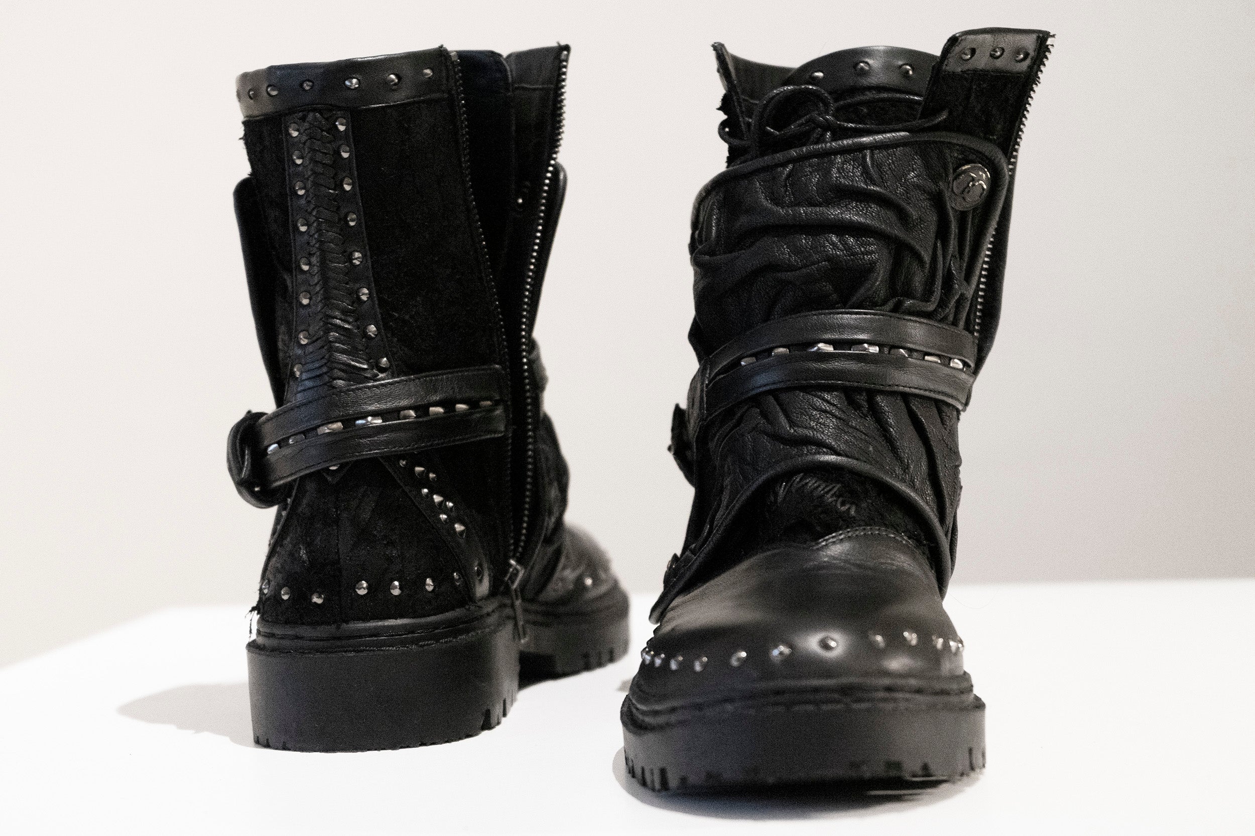 The MAGMA Wasteland Studded Leather Ankle Combat Boots by Kaya Movement are displayed on a white surface. These black leather boots feature intricate stitching, metallic details, decorative straps, and zippers, creating a rugged and stylish look reminiscent of punk rock boots. One boot faces forward while the other faces backward.
