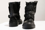 The MAGMA Wasteland Studded Leather Ankle Combat Boots by Kaya Movement are displayed on a white surface. These black leather boots feature intricate stitching, metallic details, decorative straps, and zippers, creating a rugged and stylish look reminiscent of punk rock boots. One boot faces forward while the other faces backward.