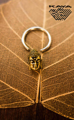 A Buddha silver ring with a gold pendant from Kaya Movement is placed on a large dried leaf with visible veins. The keychain loop is open, showcasing the intricate details of the Buddha's face. The top right corner features the "Kaya Movement" logo in a stylized font.