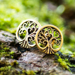 A pair of Tree of Life Stud Earrings from Kaya Movement is displayed on moss-covered bark. These earrings, crafted from elegant Yellow Brass, showcase a sophisticated gold-tone tree of life design with intricate branches and roots. The background features blurred green foliage, enhancing the natural aesthetic and making them perfect ear lobe jewelry for any setting.