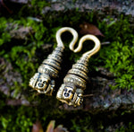 Two intricately designed ANGKOR WAT Heavy Ear Weights in Gold from Kaya Movement lie side by side on a moss-covered surface. Each 6 gauge piece features a detailed Buddha carving at the bottom, with ornate patterns leading up to a curved hook at the top. The background consists of green moss and textured bark.