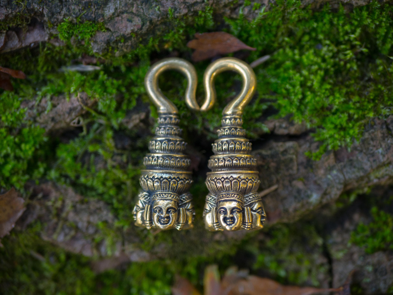 A pair of ANGKOR WAT Heavy Ear Weights in Gold from Kaya Movement rest on a bed of green moss. Each piece of 6 gauge body jewelry showcases detailed carvings, with multiple tiers and a serene Buddha face at the bottom, evoking traditional cultural artifacts. Fallen leaves are scattered around.