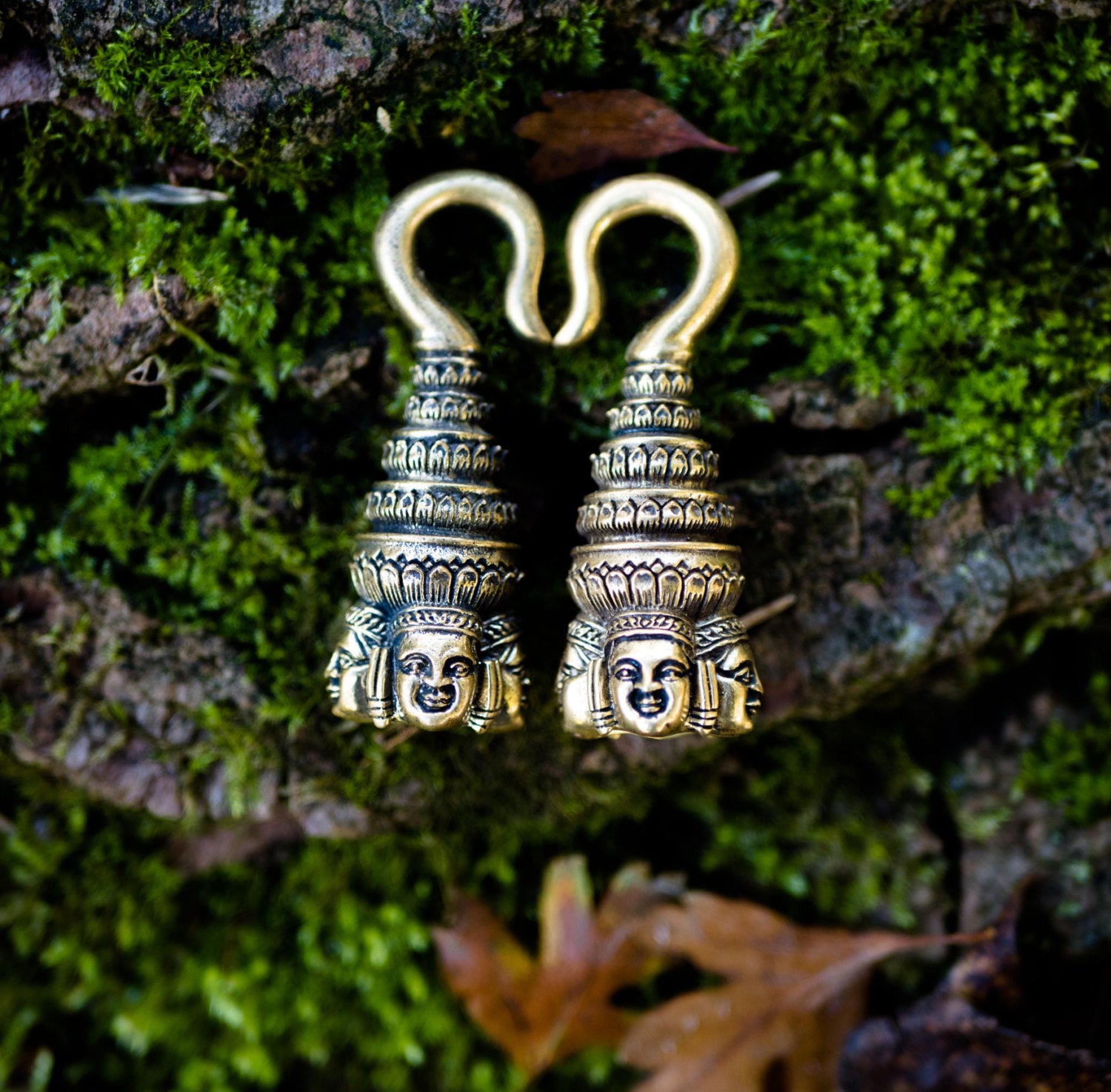 A pair of intricately designed ANGKOR WAT Heavy Ear Weights in Gold by Kaya Movement, resembling spiral towers with detailed human faces at the base, lying on a lush green mossy surface with scattered brown leaves.