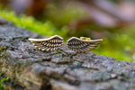 The EL Angel Wings Stud Earrings in Gold from Kaya Movement rest on a textured piece of wood surrounded by soft green moss, providing a natural and earthy backdrop. Crafted from yellow brass, these 18-gauge earrings feature intricate detailing that highlights the delicate feathers and curves of the wings, making them a perfect choice for elegant gift packaging.