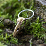 The CROW SKULL Gothic Silver Ring with Gold Charm by Kaya Movement, available in 18, 16 or 14 gauge, rests on a bed of green moss and rocks. The backdrop of natural foliage highlights the intricate craftsmanship of this handcrafted jewelry piece featuring a detailed bird skull design.