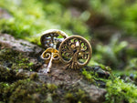 Close-up of Kaya Movement's Tree of Life Stud Earrings in Gold | 18 gauge, resting on moss-covered wood. These circular earrings, crafted with intricate cutouts representing the tree's branches and roots, are showcased in a natural, earthy setting. The yellow brass material adds a warm touch to these beautiful pieces.