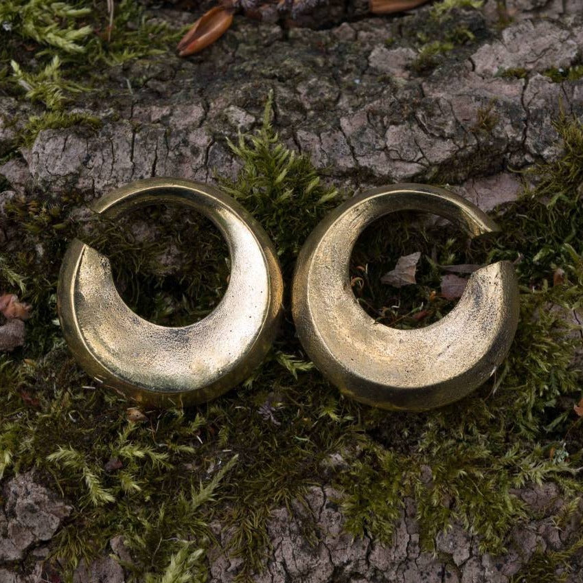 The HOLO Hoop Dangle Ear Weights in Gold from Kaya Movement, with a crescent-shaped design, are placed against a background of rough tree bark and patches of green moss. The ear weights have a shiny, textured surface, with small pieces of natural debris scattered around.