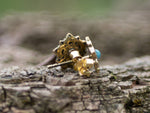 A close-up photo of the OLLY Mandala Stud Earrings in Gold with Turquoise or Onyx from Kaya Movement on a rough, textured surface, possibly wood. Crafted from high-quality yellow brass, these 18 gauge earrings feature a small blue jewel reminiscent of natural turquoise and have intricate details including a sunburst or floral design, adding an ornate touch to the jewelry.