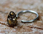A close-up photo of a silver ring laid on a textured surface. The handcrafted GANESHA Silver Ring with Gold Pendant by Kaya Movement features an intricately designed gold-colored metallic ornament resembling a lion's or dragon's head. The details on the ornament include carved elements and embellishments, showcasing expert craftsmanship. Available in 18, 16, or 14 gauge.