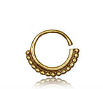 The IRI Seamless Septum Ring in Gold by Kaya Movement is a small, circular nose ring crafted with a textured design on the bottom half that features a series of small gold beads. This 16 gauge, shiny metal piece is open on one side and reflects light beautifully against a white background, highlighting its intricate craftsmanship and subtle tribal ethnic design.