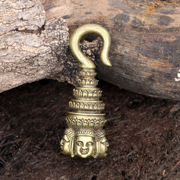 The ANGKOR WAT Heavy Ear Weights in Gold by Kaya Movement rest on a rustic wooden surface. Resembling a traditional ceremonial headdress, these intricately detailed ear weights feature a Buddha carving with multiple tiered layers rising above it. The top of the pendant forms a hook shape, perfect for brass ear weights or any body jewelry.
