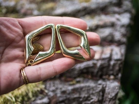 A hand adorned with gold rings holds two DAYAK Triangle Keyhole Ear Hangers in Gold (00 gauge) by Kaya Movement. The background features a textured tree trunk with moss.